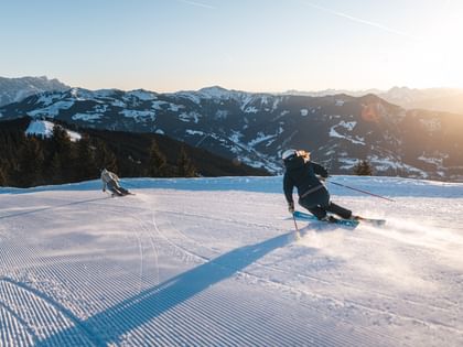 Two skiers make dynamic ski turns on a groomed slope. The low morning sun illuminates the snow-covered mountain landscape.