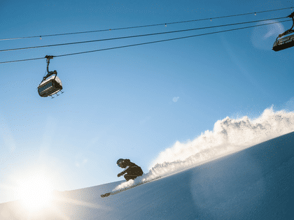 Skifahrer beim hinunterfahren auf der Piste mit einem Sessellift über ihm und schönem Wetter.