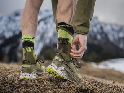 Close-up of DYNAFIT trail running shoes with green and black socks on a muddy mountain trail, with snow-covered mountains in the background.