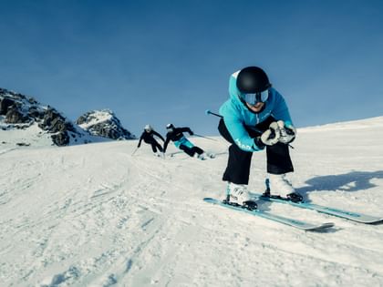 Eine Person in türkisfarbener Skijacke, schwarzem Helm und Skibrille fährt in tiefer Hocke auf HEAD Ski eine verschneite Piste hinunter. Zwei weitere Personen folgen vor felsigen Berggipfeln und blauem Himmel.
