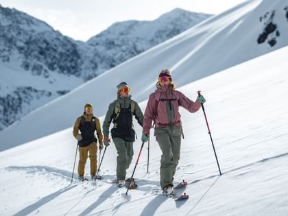 Das Bild zeigt drei Skifahrer in Winterkleidung, die mit Skistöcken und Tourenski mit Fellen einen verschneiten Berg hinaufwandern. Im Hintergrund ist eine beeindruckende Berglandschaft zu sehen.