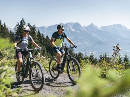 Eine Dame und ein Herr am E-Bike auf einer Schotterstraße in den Bergen. Rund herum erblüht die Natur. 