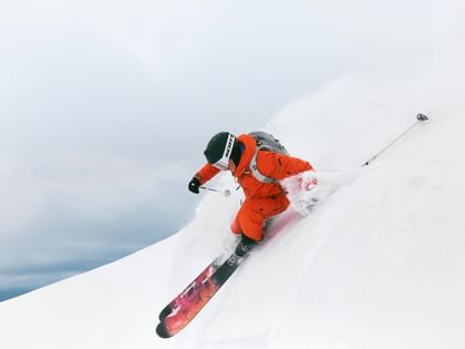 Peak Performance AW 25 A skier in orange ski gear carves powerfully through fresh powder on a steep slope. Snow sprays up as he makes a dynamic turn.