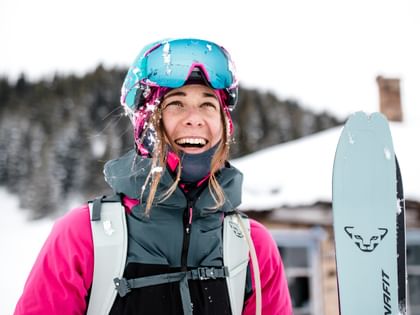 Smiling female ski tourer wearing a pink jacket, helmet and ski goggles carries a DYNAFIT touring ski in a snowy mountain landscape.