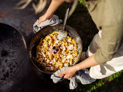 A person holds a large pan of freshly prepared Kaiserschmarrn dusted with powdered sugar by a fire pit. A spatula rests inside the pan.