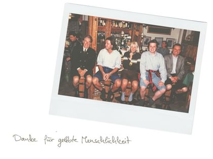 Eine Gruppe von Frauen und Männern sitzt gemeinsam in einem Restaurant und lächelt in die Kamera. Darunter steht handschriftlich „Danke für gelebte Menschlichkeit“.
