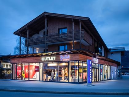Das Bild zeigt das Bründl Sports Outlet bei Dämmerung. Das Gebäude ist beleuchtet, was die "Outlet" und "Bründl Sports" Schilder gut sichtbar macht. Das Schaufenster zeigt sportliche Winterbekleidung.