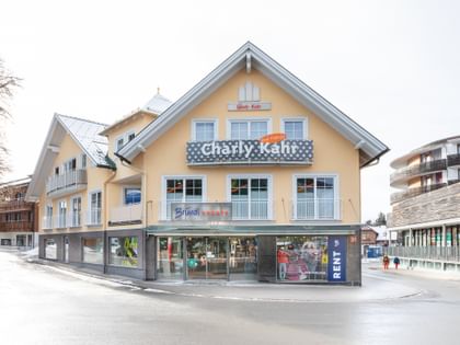 Das Bild zeigt ein Gebäude mit dem "Bründl Sports" Shop im Erdgeschoss und einem Schild für "Charly Kahr" im oberen Bereich, umgeben von anderen Gebäuden in einer winterlichen Umgebung.
