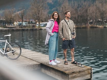 A woman wearing glasses and a man stand side by side on a wooden dock by the water, looking into the distance. On the left, a bicycle is visible, and in the background there is a calm lake, trees, and some buildings.