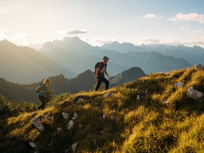 Two people with backpacks walk up a grassy slope. In the background, several mountain ranges are visible in the warm light of the low sun.