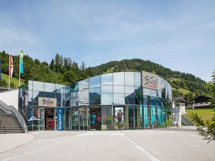 The image shows the Bründl Sports AreitXpress valley station shop with a large glass facade, shop entrance, and visible “Bründl Sports” lettering on the building. Flags and stairs are on the left, with forest, meadows, and a mountain in the background; the text “SKIDEPOT” is also visible on the building.