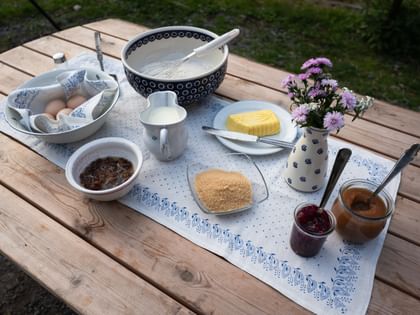 Ingredients for Kaiserschmarrn are arranged on a wooden table, including eggs, flour, milk, butter, sugar and raisins, along with jars of fruit compote. A vase with purple flowers stands beside them.