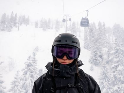 Eine Skifahrerin mit schwarzem Skihelm, violetter Skibrille und schwarzer Skijacke steht vor einer tief verschneiten Winterlandschaft. Im Hintergrund sind schneebedeckte Bäume und mehrere Gondeln einer Seilbahn zu sehen.