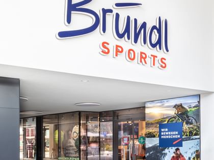 Der Eingang eines Bründl Sports Shops mit großem Markenlogo über der Glasfront. Auf einer Werbetafel sind Sportmotive und der Text „WIR BEWEGEN MENSCHEN“ zu sehen.