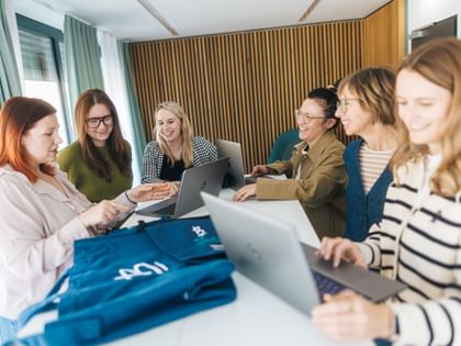 Mehrere Frauen stehen und sitzen an einem Tisch mit geöffneten Laptops und betrachten blaue Kleidungsstücke. Eine Frau zeigt auf ein Detail, während die anderen lächeln und auf die Kleidung schauen.