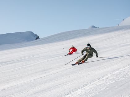 Zwei Skifahrer in roten und grünen Anzügen fahren nebeneinander die Piste hinunter, beide in dynamischer Carving-Haltung.