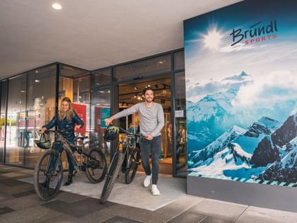 Two cyclists with mountain bikes in front of a Bründl sports shop with a large advertising poster of a snow-covered mountain landscape.