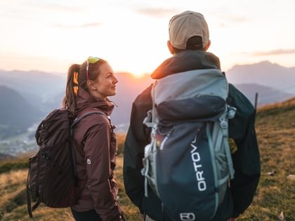 Eine Frau und ein Mann mit Rucksäcken stehen auf einem grasbewachsenen Hügel und schauen in Richtung der aufgehenden Sonne. Im Hintergrund sind Bergketten und ein Tal zu sehen, während warmes Licht die Szene beleuchtet.