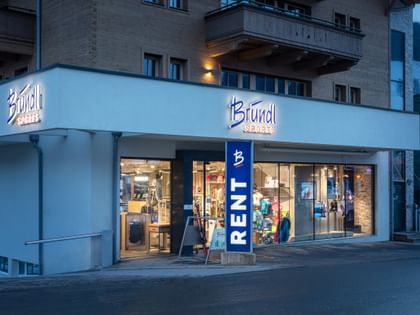Das Bild zeigt den Eingang eines Bründl Sports Shops in einem alpinen Gebäude. Über dem Eingang befindet sich das Logo, und der Schriftzug "RENT" weist auf die Verleihmöglichkeit hin.
