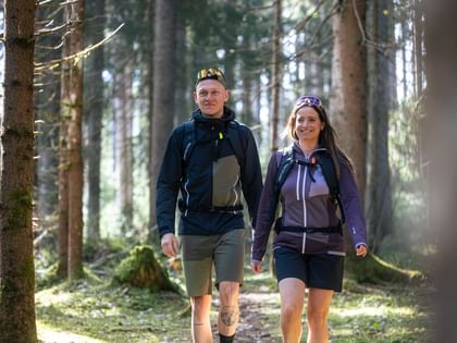 Two hikers are enjoying a sunny day in the forest, wearing functional outdoor clothing and backpacks.