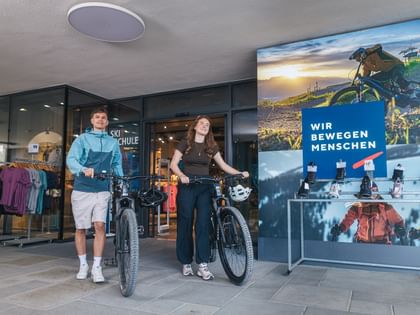 Zwei Radfahrer mit Mountainbikes vor einem Bründl Sportgeschäft mit großem Werbeplakat einer verschneiten Berglandschaft.