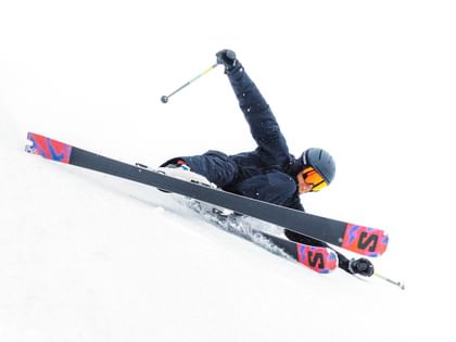 Ein Skifahrer in schwarzer Ausrüstung legt sich dynamisch in die Kurve auf einer schneebedeckten Piste. Der Fahrer trägt einen schwarzen Helm und orange Skibrille, während Schnee unter den Kanten aufspritzt.