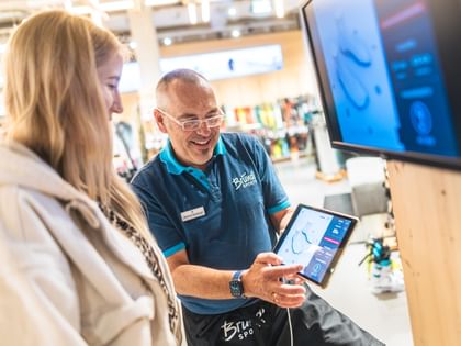 Bootfitting Ein Bründl Sports Mitarbeiter berät eine Kundin im Shop und zeigt ihr mithilfe eines Tablets eine digitale Analyse für Skischuhe. Im Hintergrund sind Sportartikel und Skiausrüstung zu sehen.