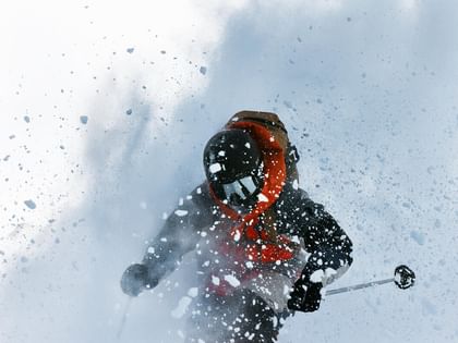 A skier wearing a dark jacket with an orange hood rides through deep powder snow. Snow swirls intensely around, partially obscuring the view.