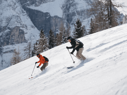 Two skiers descending through fresh powder snow in alpine terrain, wearing functional Martini ski touring apparel.