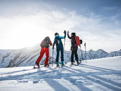 Three people in ski touring gear stand on a snowy ridge, with two of them happily giving each other a high-five, surrounded by an impressive mountain landscape.