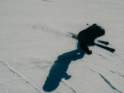 Eine Skifahrerin in schwarzer Skibekleidung und Helm fährt in tiefer Haltung über eine verschneite Piste. Die Aufnahme aus der Vogelperspektive zeigt aufgewirbelten Schnee und einen langen Schatten auf der Piste.