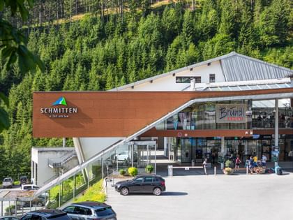 Das Gebäude hat eine auffällige Architektur mit einer großen Glasfassade und einem markanten, schrägen Dach. Vor dem Eingang sind einige Autos und Menschen zu sehen, die das Geschäft betreten oder verlassen. Über dem Eingang befindet sich das Bründl Sports-Logo, und daneben ist das Schmitten-Logo auf der braunen Fassade angebracht. Die Umgebung ist von dichten Wäldern umgeben, die eine idyllische Kulisse bilden.