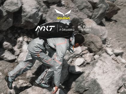 Ein Bergsteiger in grauer Funktionskleidung mit orangefarbenen Akzenten klettert über felsiges Gelände. Er trägt einen schwarzen Rucksack und bewegt sich konzentriert nach vorne.