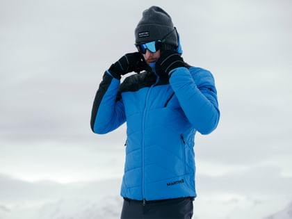 The image shows a man in a blue winter jacket preparing himself on a snow-covered mountain and adjusting his gloves.