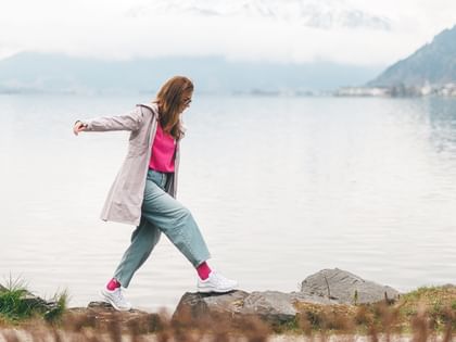 A woman wearing glasses carefully walks across large rocks at the edge of a lake, with her arms slightly outstretched. She is wearing a pink top, a light jacket, and jeans, with calm water and misty mountains in the background.