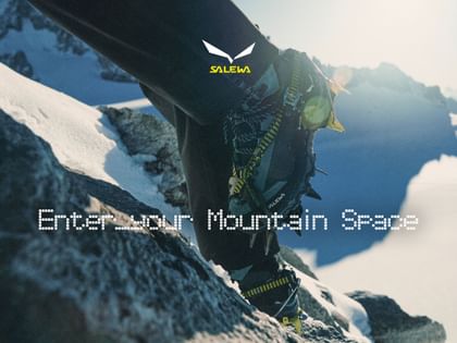 Close-up of mountaineering boots fitted with crampons while climbing over snow-covered rocks, with sunlit mountain peaks in the background. The image features the Salewa logo and the text “Enter your Mountain Space”.