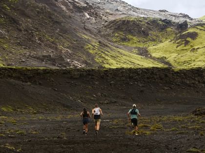 Drei Läufer rennen durch eine weite vulkanische Berglandschaft mit steilen Hängen und moosbewachsenen Felsen.