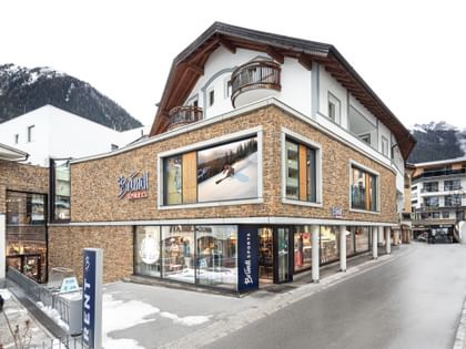 Bründl Sports Ischgl Zentrum mit Steinfassade, großen Schaufenstern und Bründl Sports Logos bei Tageslicht. Vor dem Shop steht ein „RENT“-Schild; Schneereste säumen die Straße und schneebedeckte Berge liegen im Hintergrund.