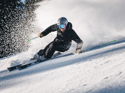 A skier in black ski clothing carves low across the slope, creating a large cloud of snow.