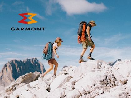 Two people with large backpacks walk across bright rocks on a mountain. In the background, more mountain peaks and a blue sky with a few clouds are visible.