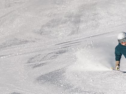 A skier wearing a teal ski outfit, a white helmet and goggles makes a sharp turn on a groomed slope. Snow sprays up behind the skis.