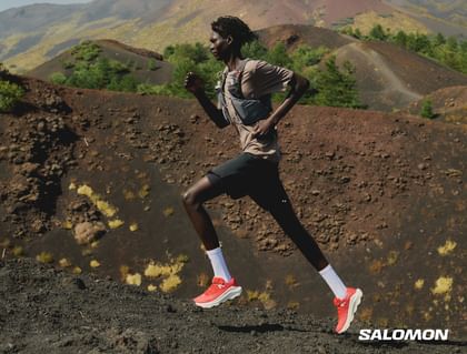 A trail runner runs across dark volcanic terrain in a mountainous landscape while wearing Salomon trail running shoes and a running vest.