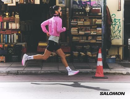 A runner wearing a pink running jacket and Salomon running shoes runs along a city street in front of a shop filled with books and vintage items.