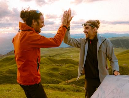Zwei Männer in Outdoorbekleidung geben sich ein High-Five auf einem Berggipfel bei Sonnenuntergang. Sie lachen, tragen Funktionsjacken und genießen den Moment nach dem Aufbau eines Zelts.