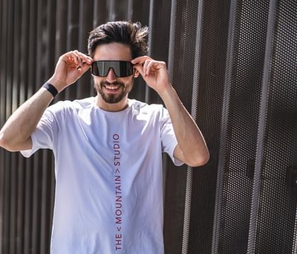 Male model in front of building, wearing white shirt and beige pants and puts his sunglasses on.