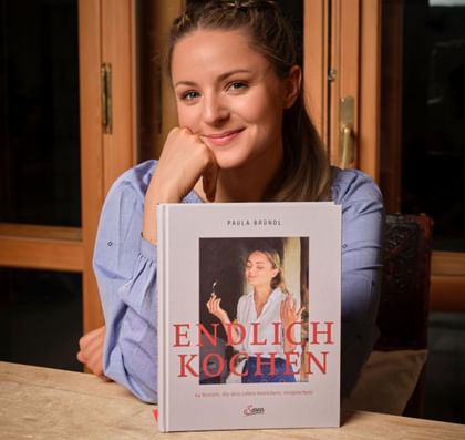 Paula am Tisch sitzend mit ihrem Kochbuch "Endlich Kochen"