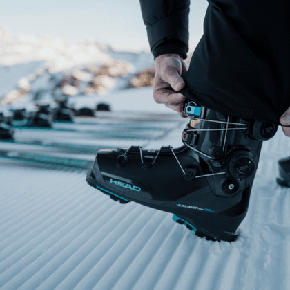 Nahaufnahme eines schwarzen HEAD Skischuhs mit türkisfarbenen Details und BOA-Drehverschlüssen auf präpariertem Schnee. Eine Person zieht das Hosenbein über den Schaft, im Hintergrund liegen mehrere Ski.