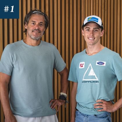 Das Bild zeigt Stefan Rettenegger (rechts) und Stefan Steinacher (links). Rettenegger, der jüngere der beiden, trägt ein hellblaues Shirt mit Logos und eine Mütze. Stefan Steinacher, links, trägt ebenfalls ein hellblaues Shirt und hat eine entspannte Haltung. Beide stehen vor einem Hintergrund aus Holzpaneelen, und oben links ist die Nummer "#1" zu sehen.