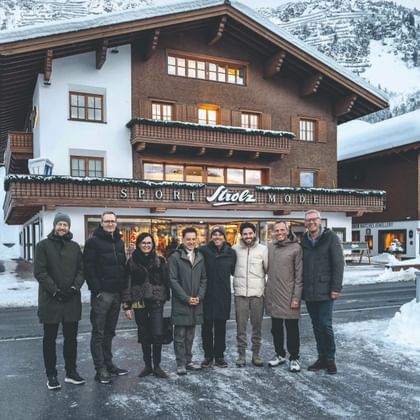 Eight people stand outside the “Sport Strolz Mode” store on a snowy street. A wooden building and snow-covered mountains are visible behind them.