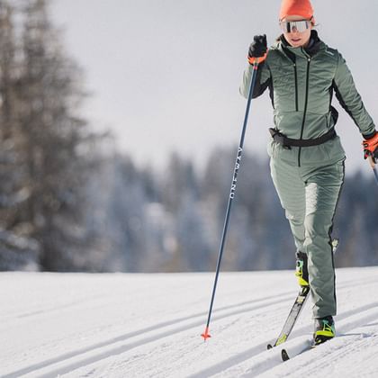 Cross-Country Skiing with Löffler – Staying Active in Winter Two cross-country skiers wearing Löffler winter sportswear glide dynamically through a snowy landscape in the sunshine.